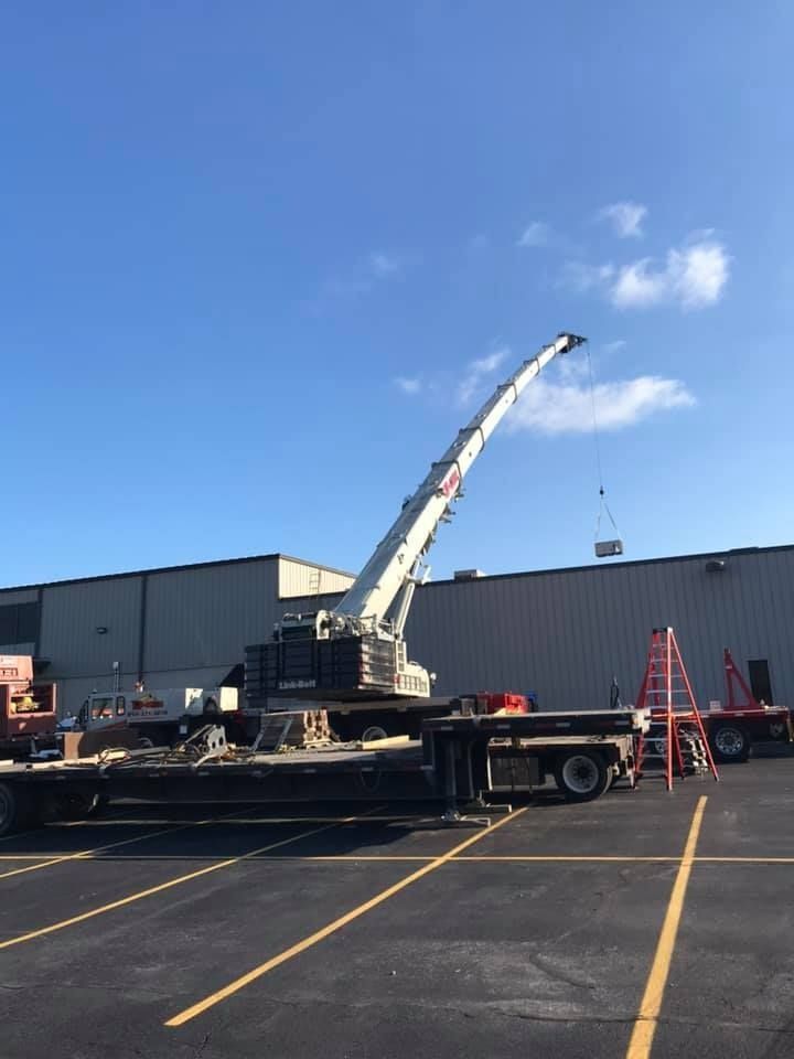 Crane lifting an object onto a building roof. Crane is white, set on a flatbed. Clear blue sky.