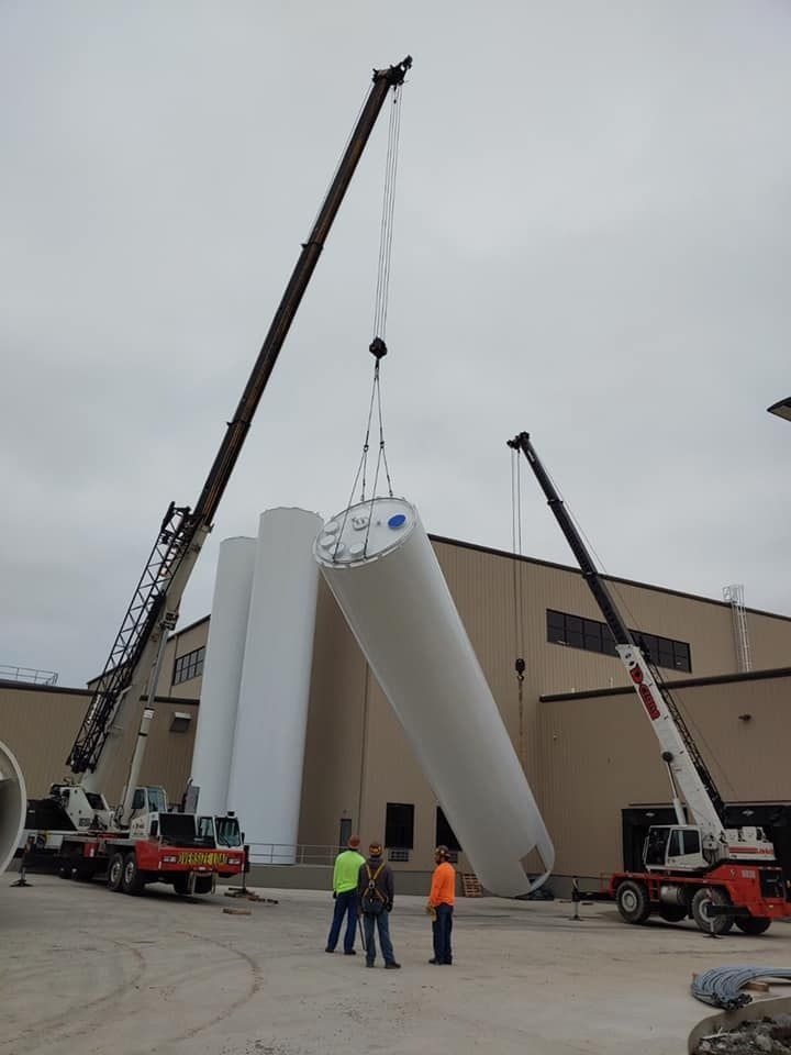 Two cranes lifting cylindrical white tanks near a building; three workers observe.