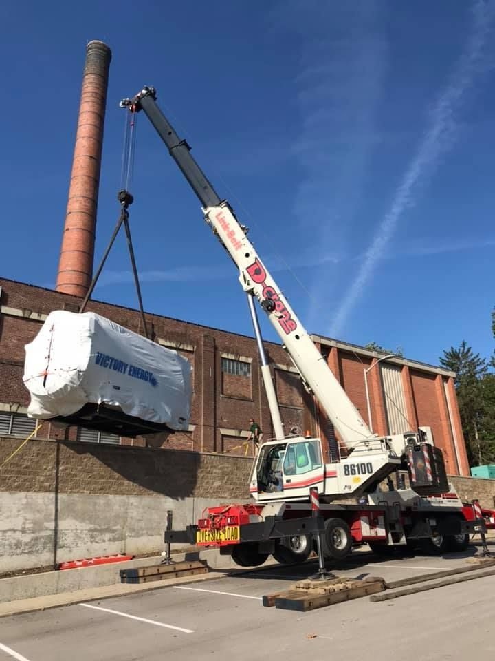 A crane lifts a large, wrapped object near a brick building and a smokestack on a sunny day.