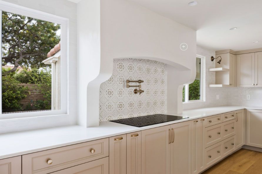 Bright white kitchen with light wood cabinets, black sink, patterned backsplash, and large windows