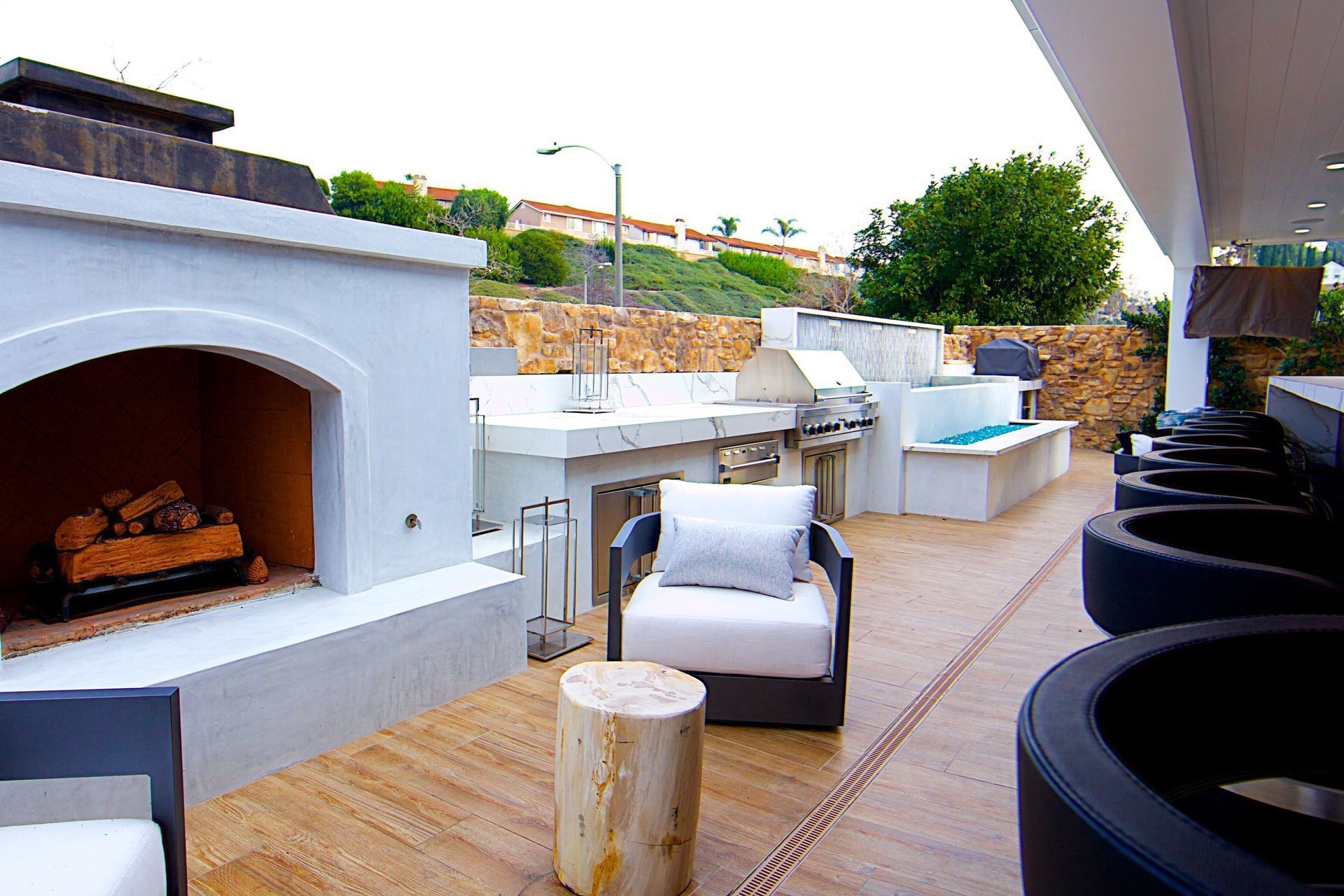 A bright, modern outdoor patio with a stone fireplace, lounge chair, grill area, and a row of black bucket-style seats.