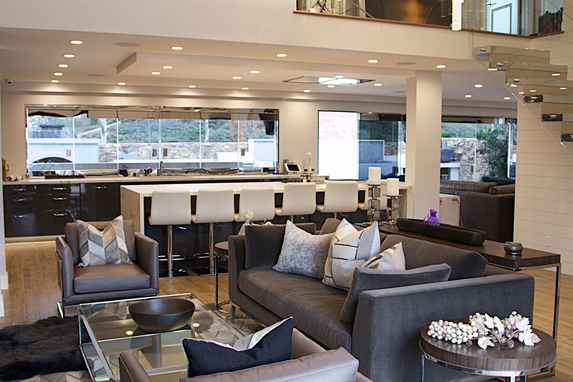 Modern, open-plan living room with grey sofas, armchairs, a glass coffee table, and a kitchen island with stools.