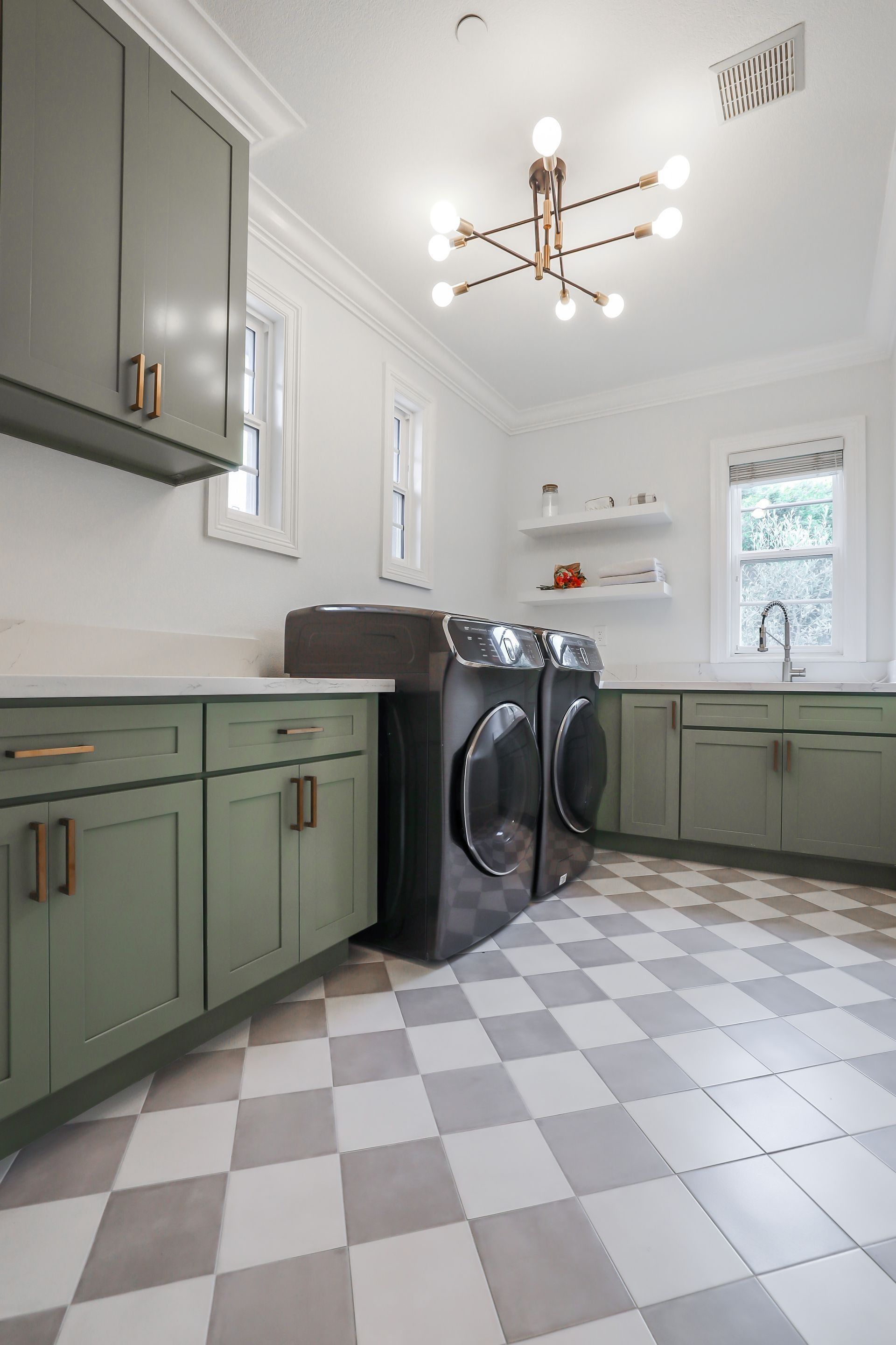 A bright laundry room with sage green cabinets, black appliances, and a gray and white checkered tile floor.