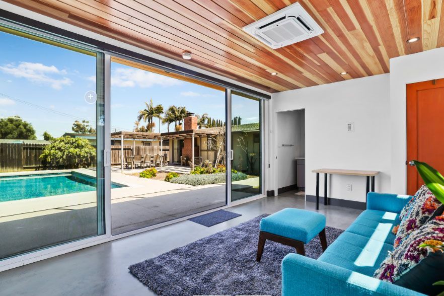 Modern living room with blue sofa, pool view through sliding glass doors, and wood-paneled ceiling
