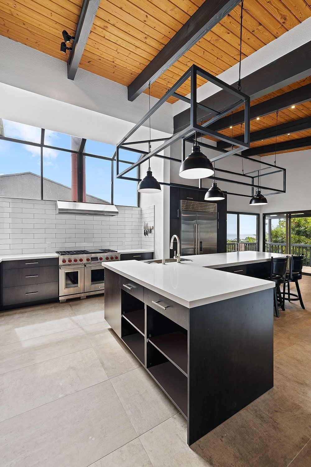 A modern, open-concept kitchen with a large central island, black cabinetry, white countertops, and wood-paneled ceilings.