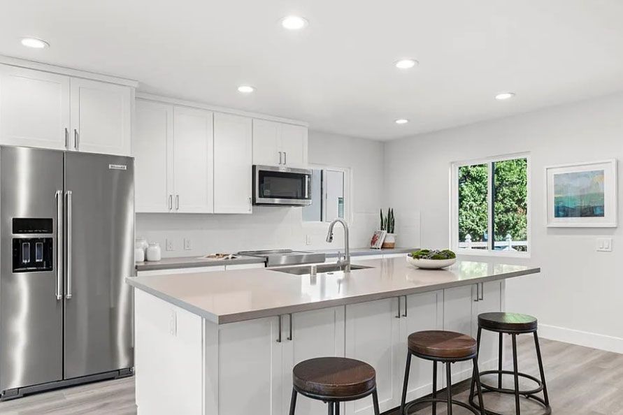 Modern white kitchen with stainless steel appliances, island seating, and a window view of greenery