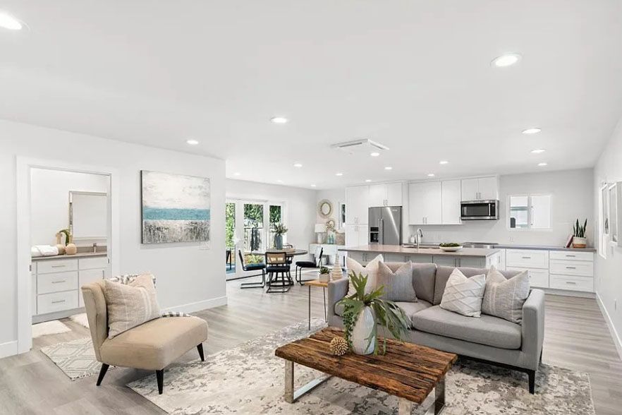 Bright open-concept living room with gray sofa, beige chair, wooden coffee table, and white kitchen in back