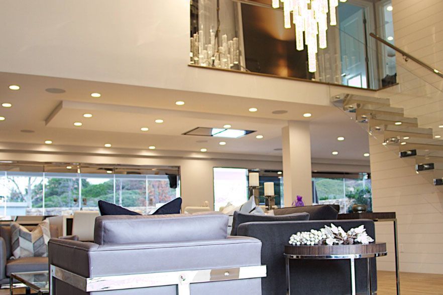 Modern living room with sofas, staircase, and large windows under a high ceiling