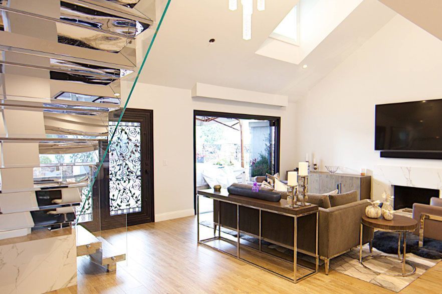 Bright modern living room with open staircase, sofa, desk, wall TV, and large windows