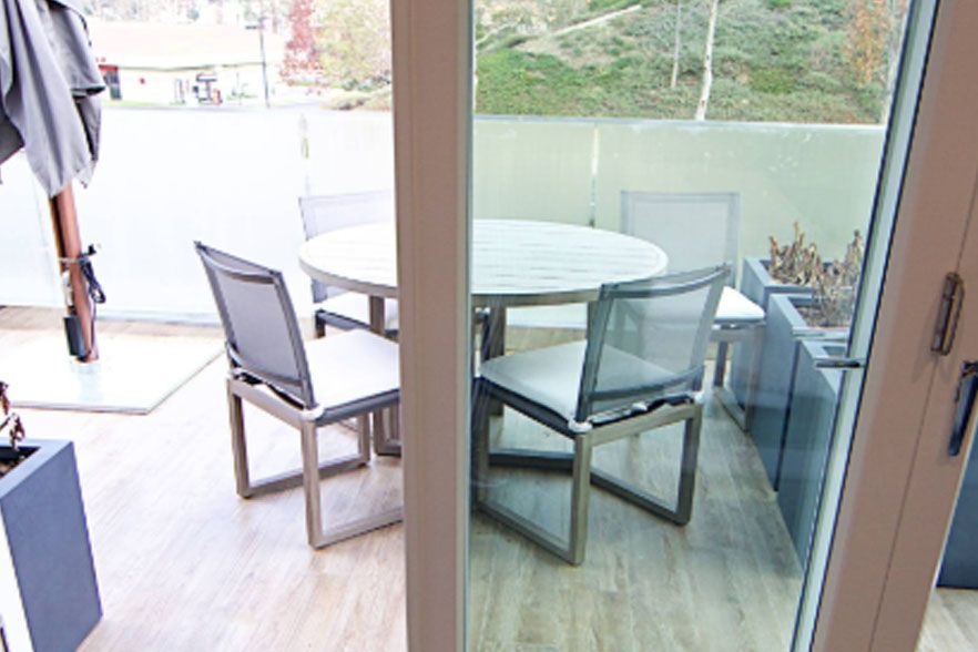 Patio with glass table and four chairs beside a sliding glass door, overlooking greenery