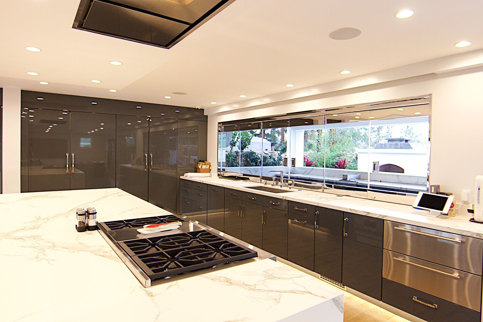 Modern kitchen featuring a white marble island with a gas cooktop, dark cabinets, and a long window overlooking greenery.
