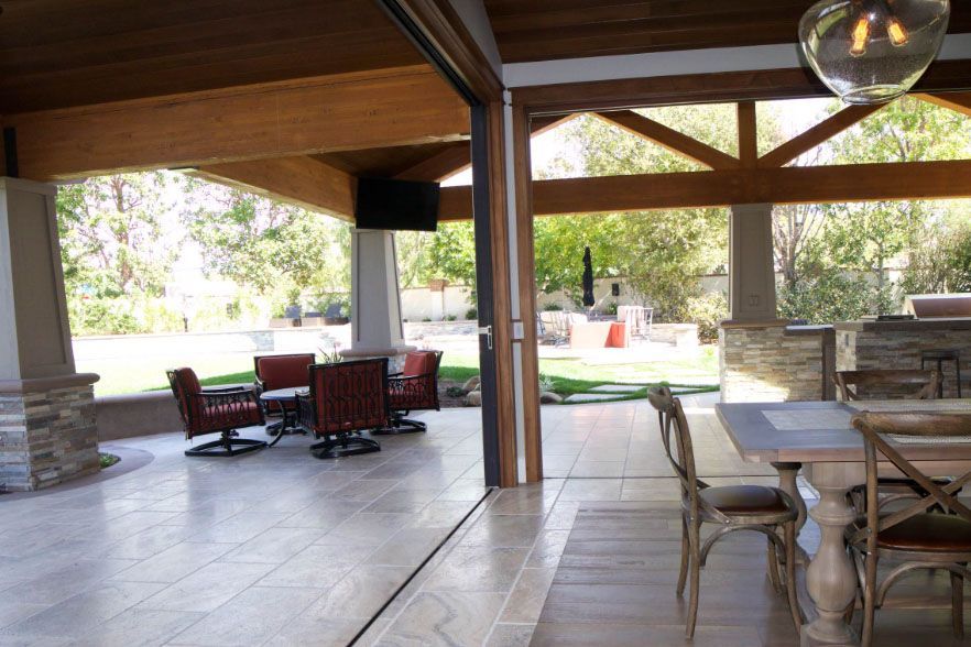 Covered patio with stone floor, wooden beams, and outdoor dining tables overlooking a pool and garden.