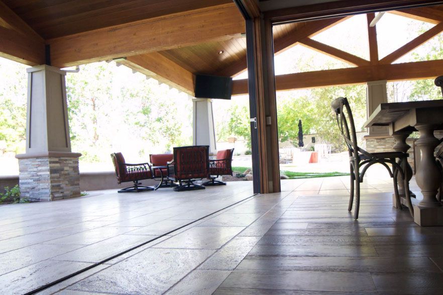 Covered outdoor patio with wooden beams, tiled floor, and wicker chairs overlooking a bright backyard
