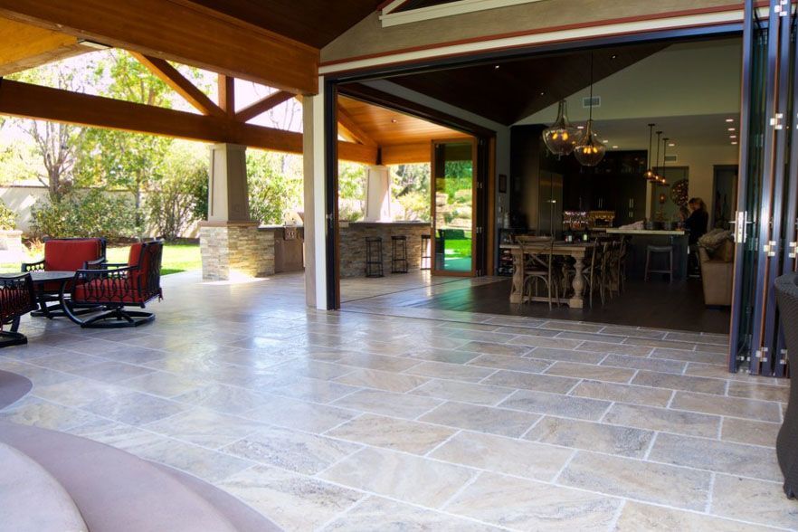 Covered patio with stone flooring, wooden beams, and outdoor seating leading to an open kitchen area
