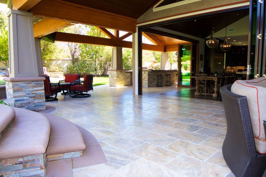 Covered patio with stone tile floor, cushioned seating, wooden beams, and open view to a sunny backyard