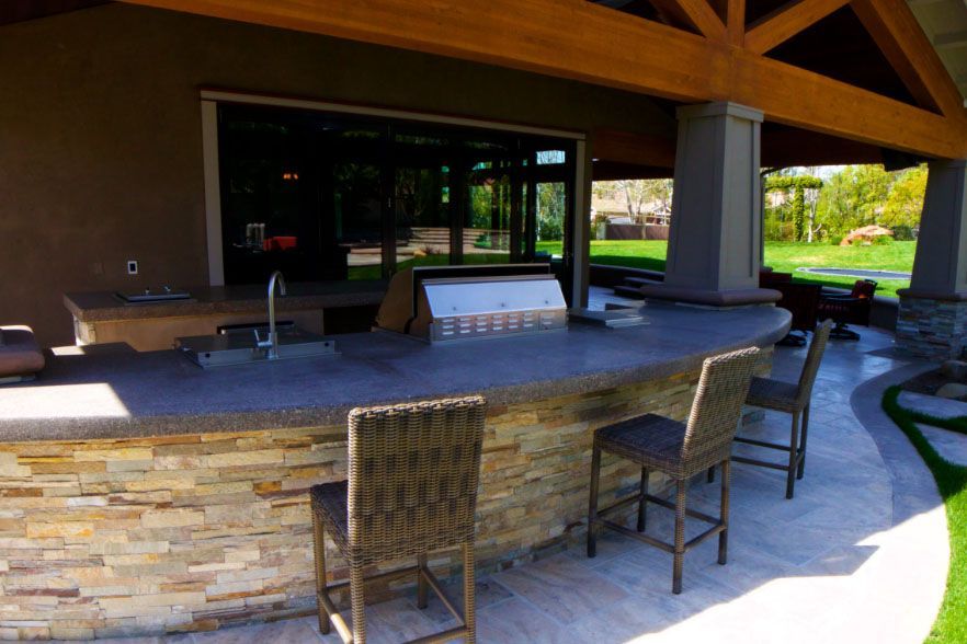 Covered outdoor bar with stone counter and woven chairs on a patio beside a green lawn