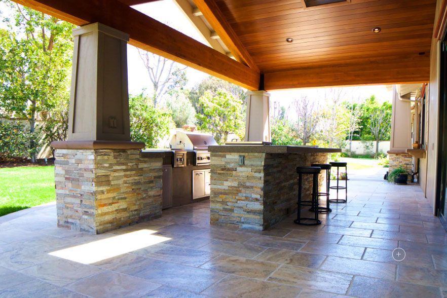 Covered outdoor patio with stone columns, wood ceiling, built-in grill, and a bar counter on tiled flooring