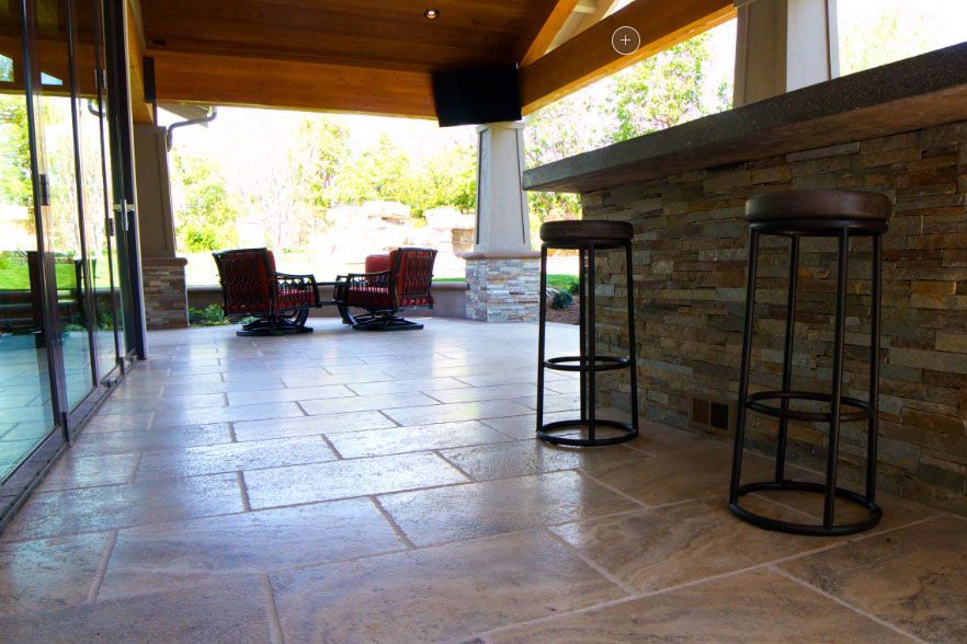 Covered patio with stone floor, bar stools, and lounge chairs overlooking a bright outdoor view