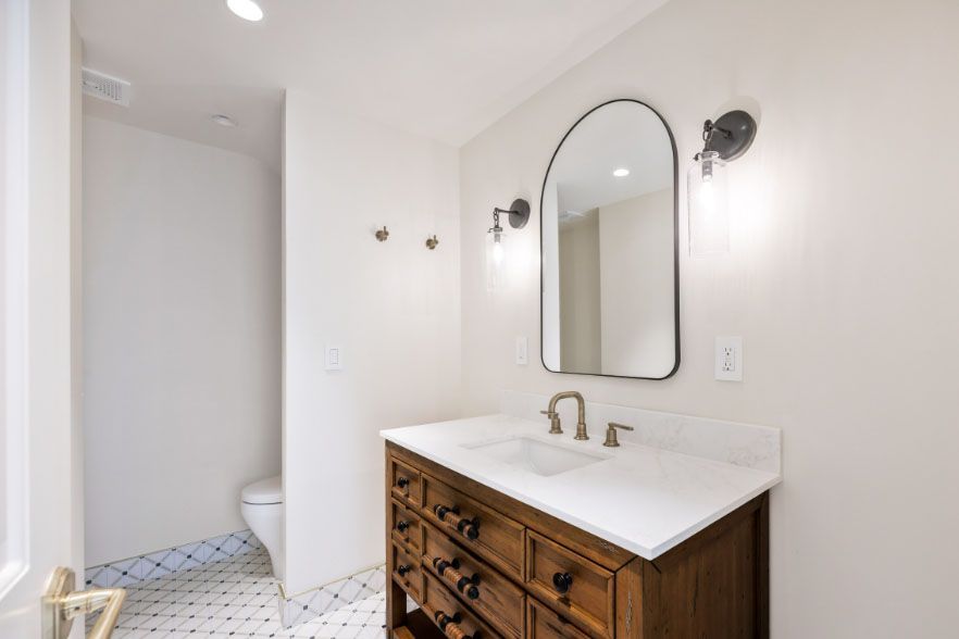 Bright bathroom vanity with arched mirror, wall sconces, and a wooden cabinet beside a tiled floor.
