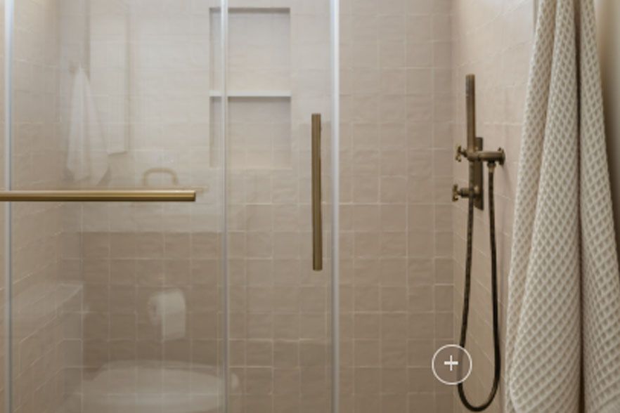 Beige tiled shower with glass door, brass grab bar, and handheld showerhead in a bathroom.