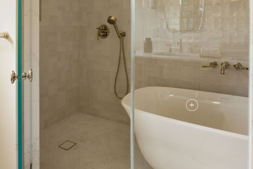 Glass shower enclosure beside a white freestanding bathtub in a beige bathroom