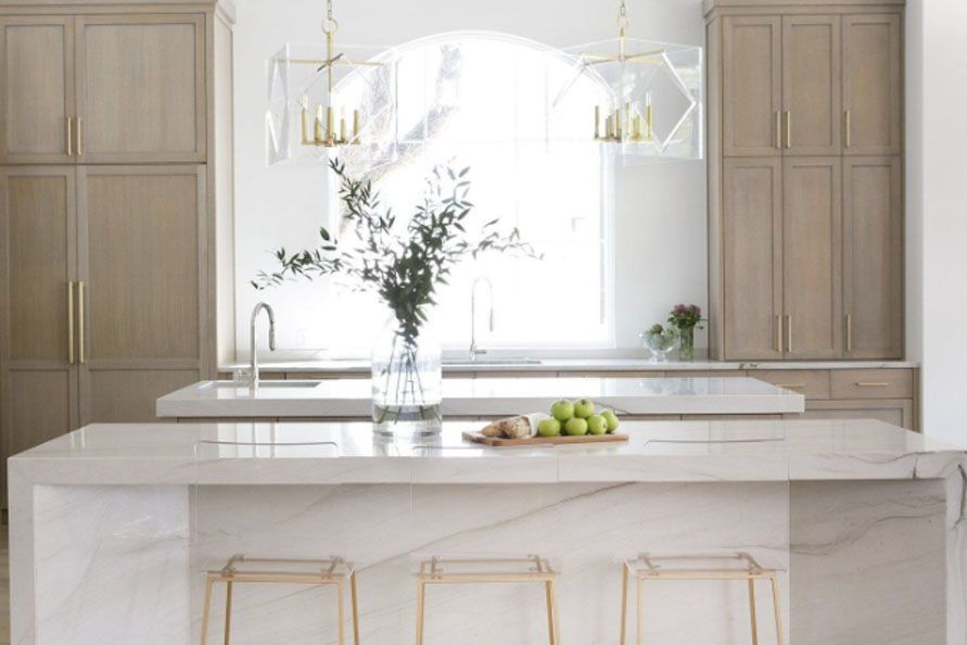 Bright modern kitchen with marble island, brass stools, wood cabinets, and a vase of flowers under pendant lights