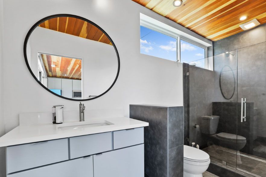 Modern bathroom with white vanity, round mirror, and glass shower under a wood-slat ceiling
