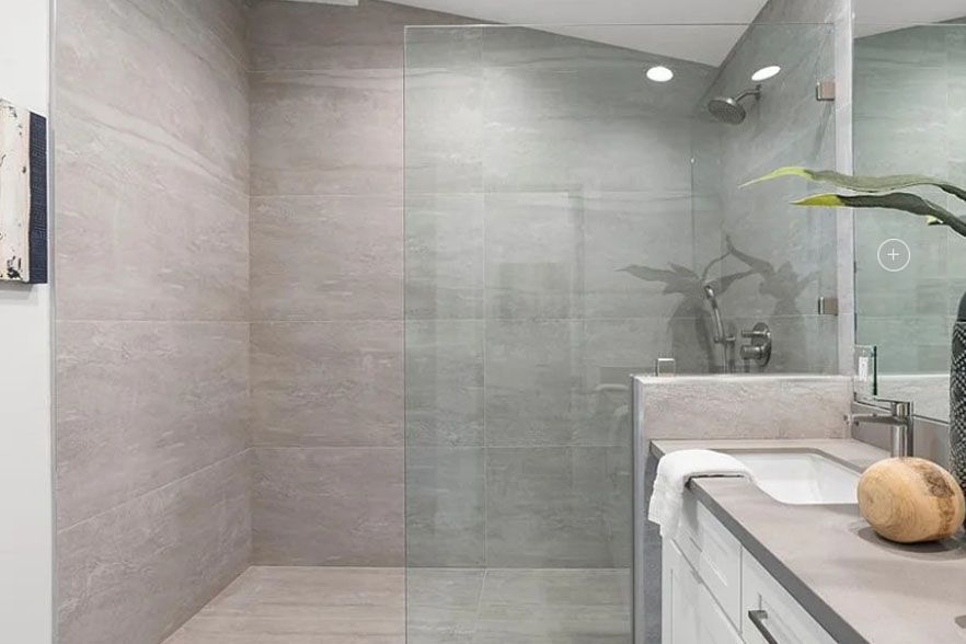 Modern bathroom with glass shower, gray tiled walls, and a white vanity with a plant and decorative bowl