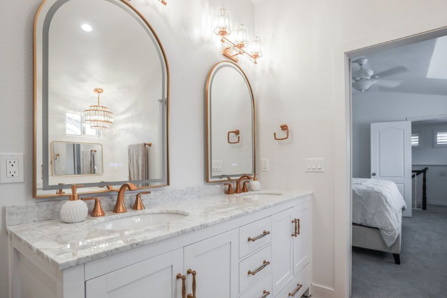 Elegant white marble bathroom vanity with gold accents and arched mirrors, opening to a bedroom view