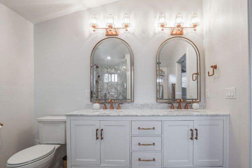 White bathroom vanity with two arched mirrors, brass sconces, and a toilet at left