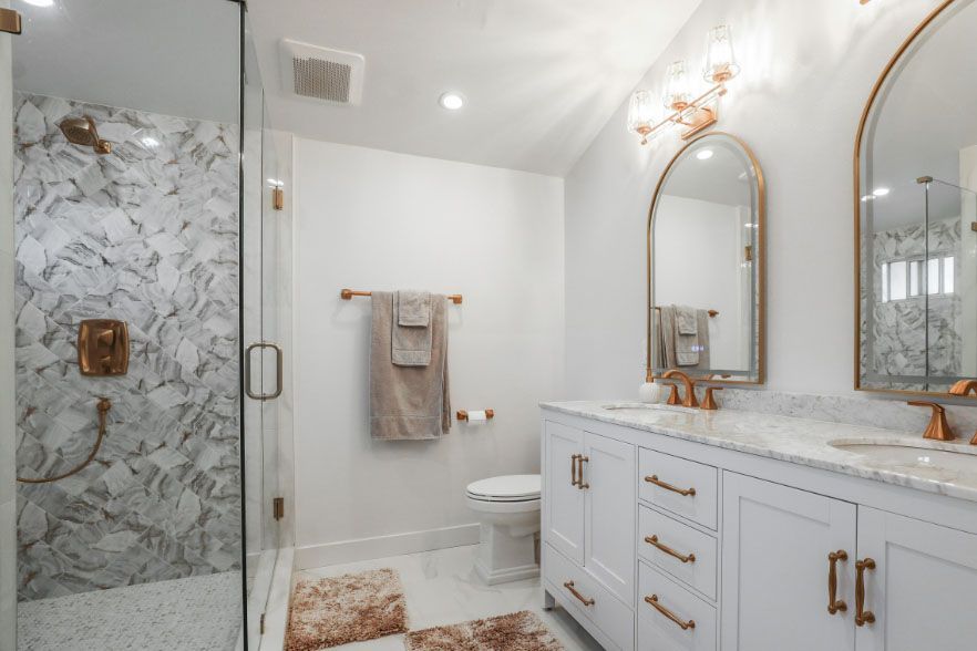 Bright white bathroom with marble shower, gold-framed mirrors, and double vanity.
