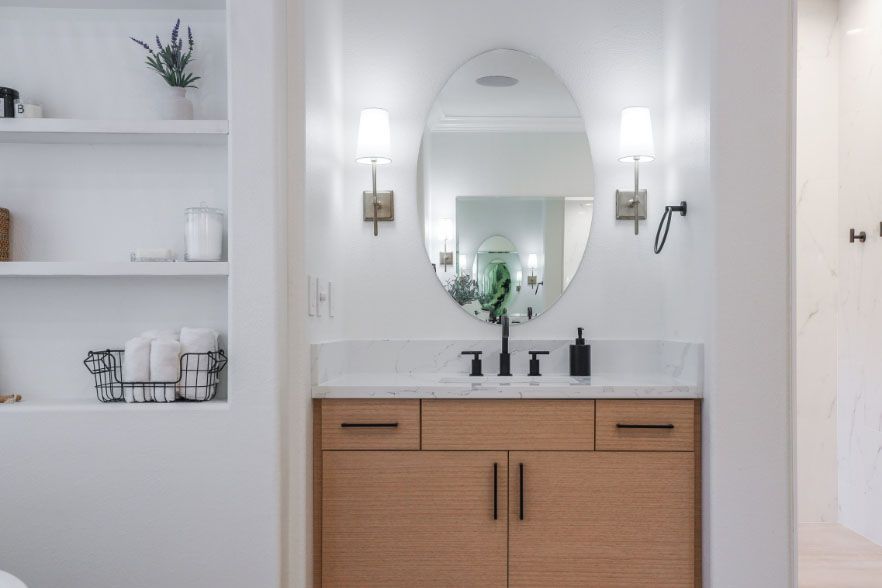 Minimal white bathroom vanity with oval mirror, brass sconces, and light wood cabinet