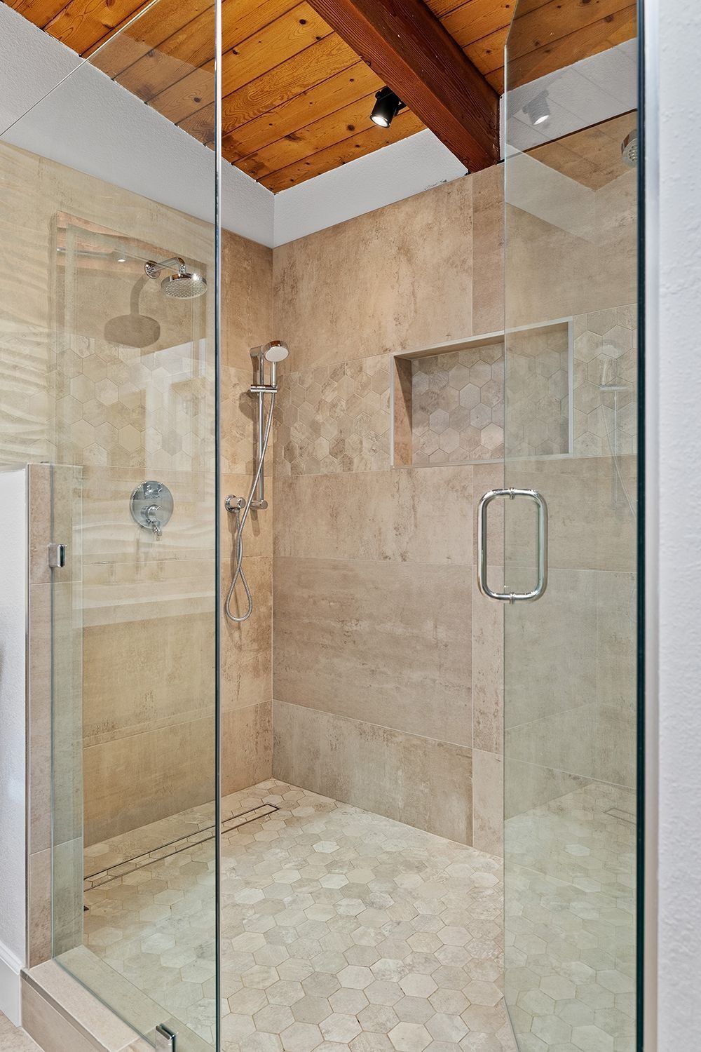 A walk-in shower with stone tile walls, a pebble floor, glass doors, and a rustic wood ceiling with a spotlight.
