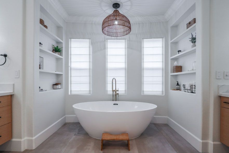 Bright white bathroom with a freestanding tub, three windows, built-in shelves, and a woven pendant light.