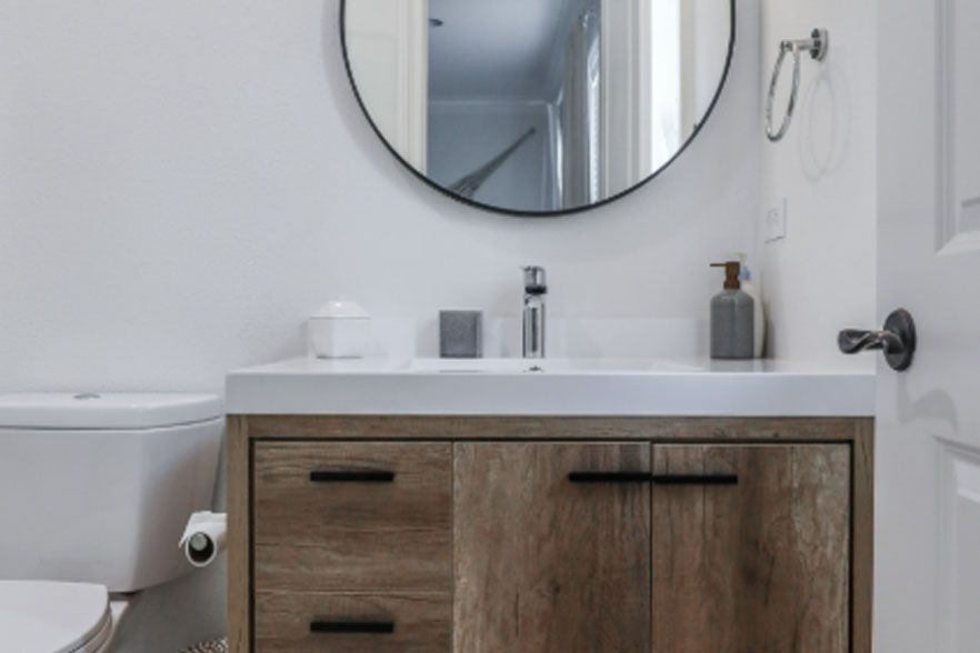 Modern bathroom vanity with round mirror, white sink, and toilet beside wood cabinet.