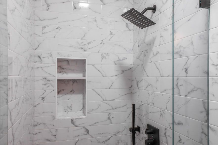 White marble-tiled shower with black fixtures, glass door, and built-in wall niche.