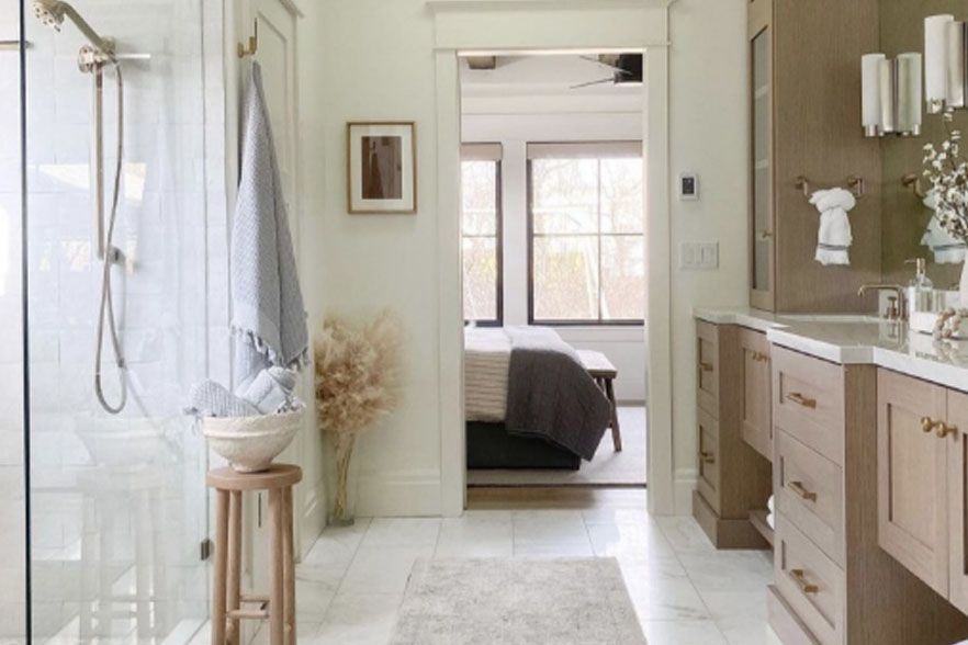 Bright, airy bathroom with glass shower, double vanity, and doorway to a bedroom beyond.