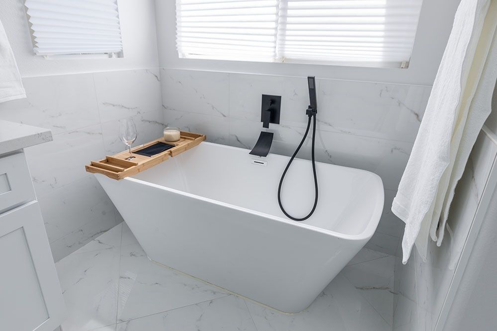 A modern white freestanding bathtub in a marble bathroom with black hardware and a wooden tray.