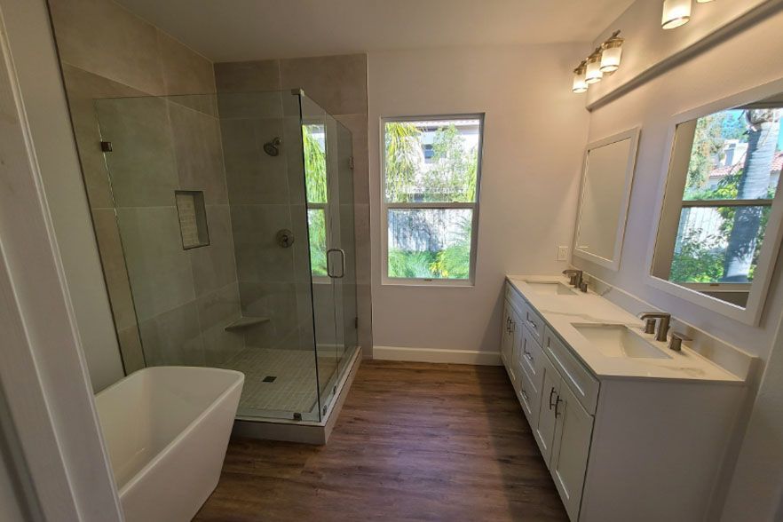 Modern bathroom with glass shower, white vanity, bathtub, and wood-look floor.