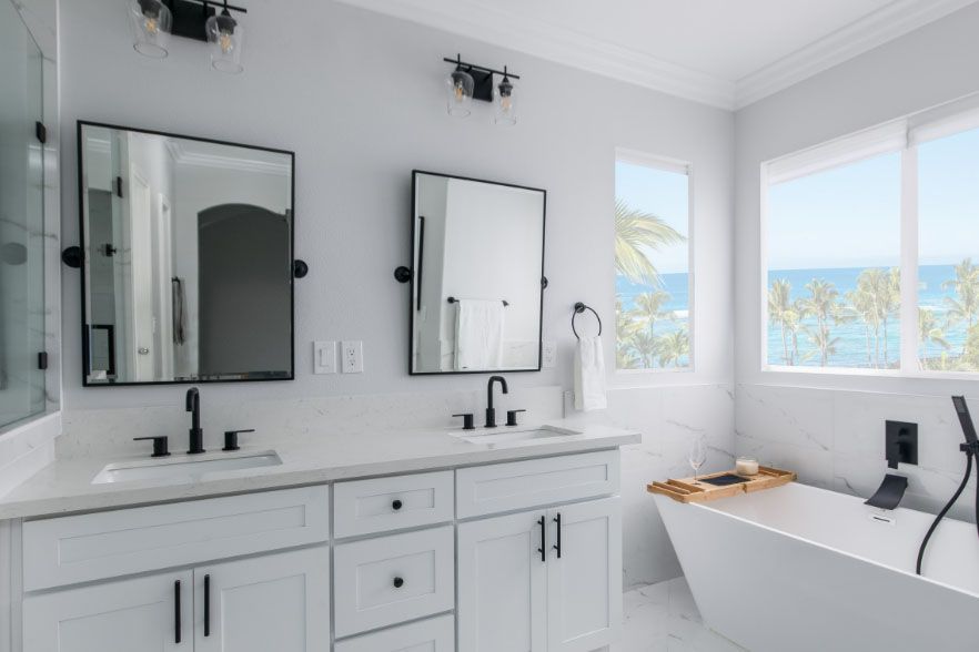 Bright modern bathroom with double vanity, mirrors, and a bathtub by large ocean-view windows.