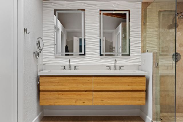 A modern bathroom vanity with a light wood floating cabinet, double sinks, two rectangular mirrors, and a wavy wall tile.