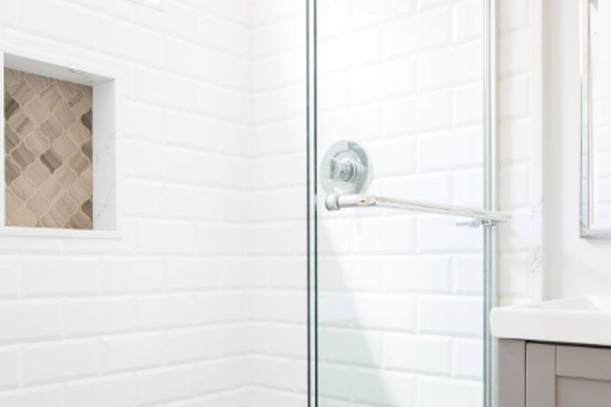 Bright white tiled shower with glass door, built-in niche, and chrome grab bar in a bathroom.