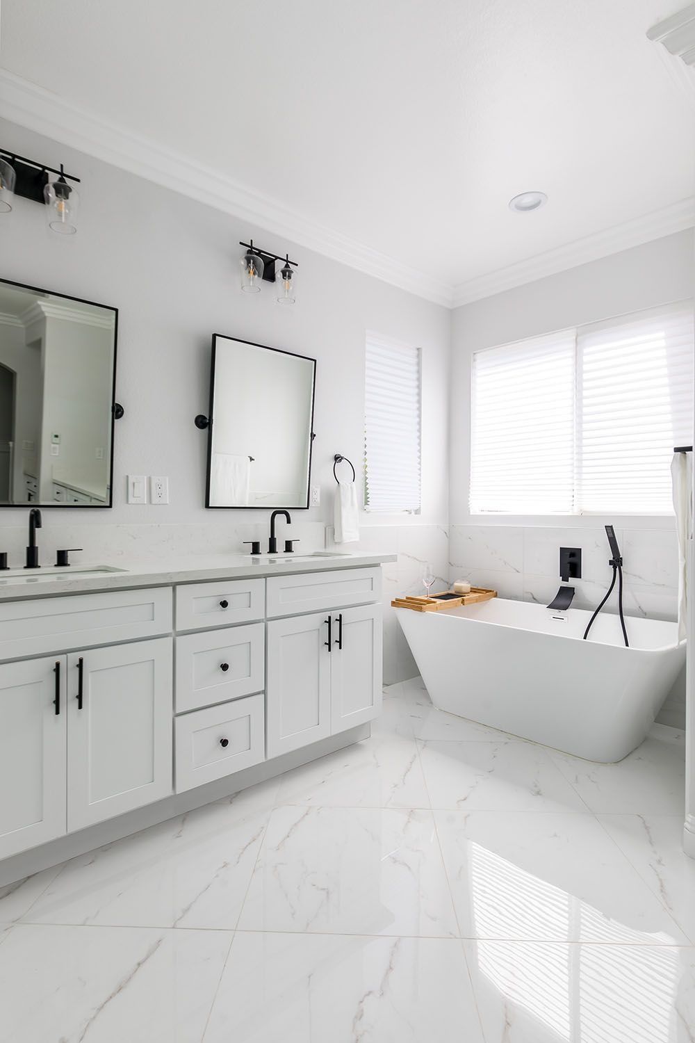 Modern white bathroom with double vanity, black fixtures, mirrors, and a freestanding tub against a large window.