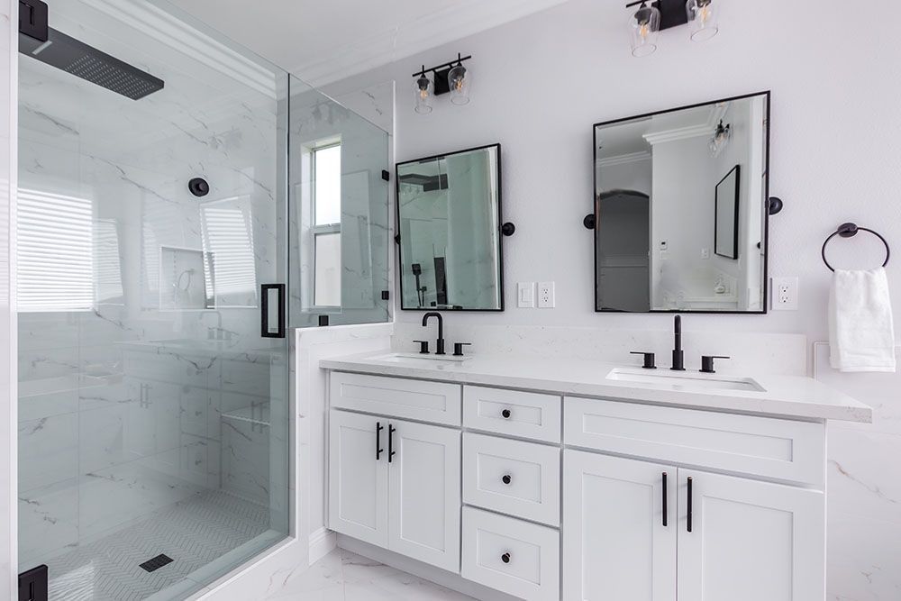 A bright, modern white bathroom featuring a marble-tiled walk-in shower, dual vanity with black hardware, and two mirrors.