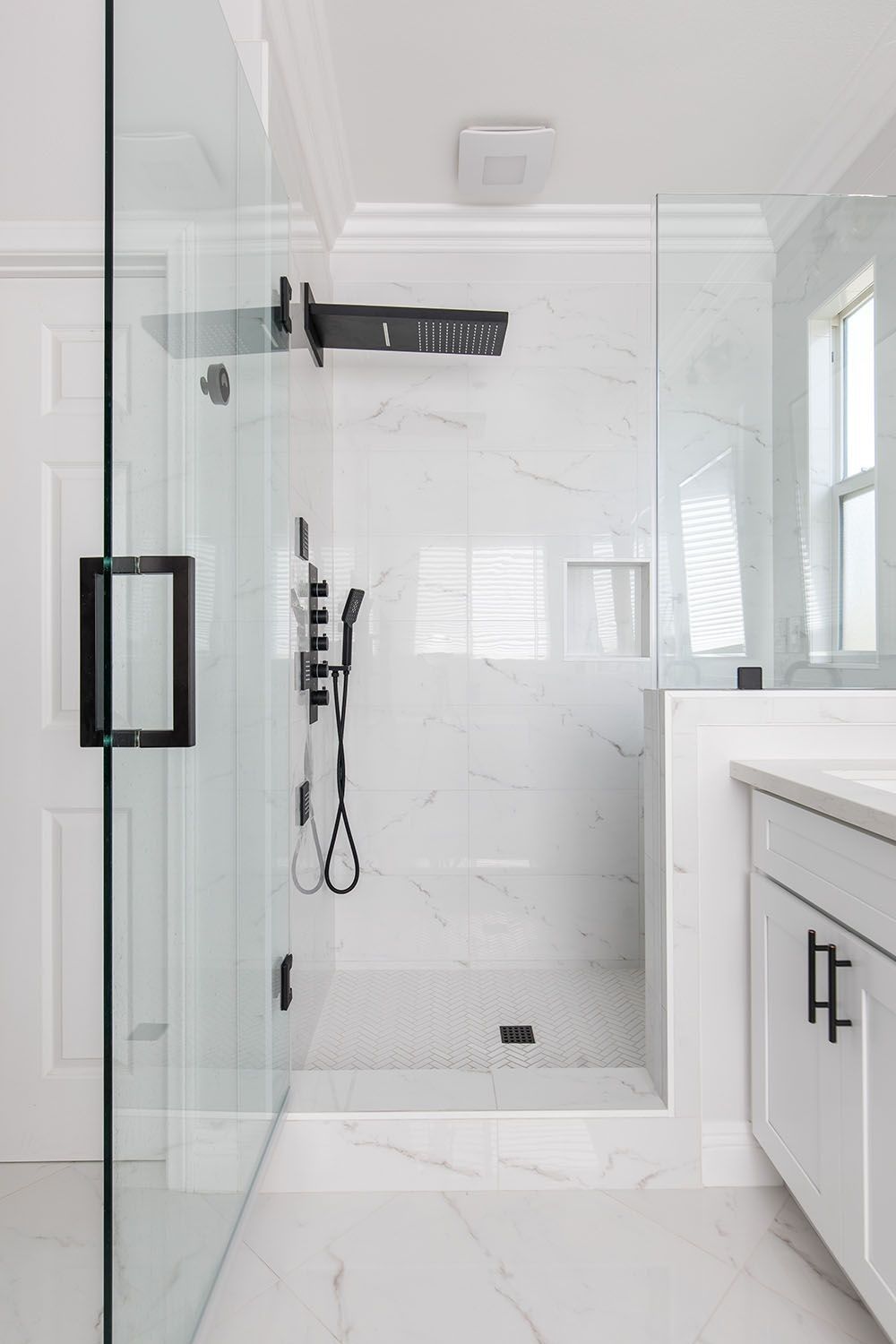 A modern bathroom with a glass-enclosed shower, white marble-patterned tiles, black fixtures, and a white vanity.
