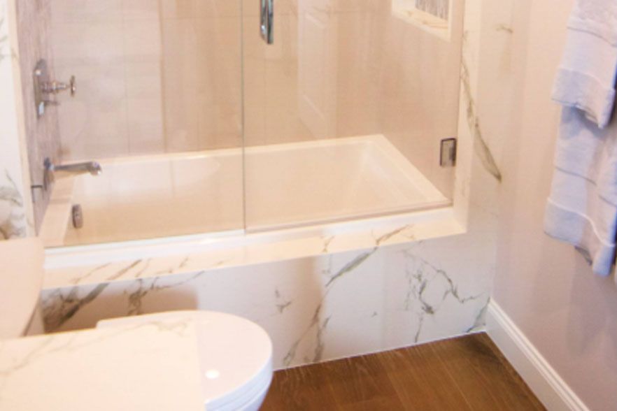 Modern bathroom with a glass-enclosed bathtub, marble tile, white toilet, and wood-look floor.