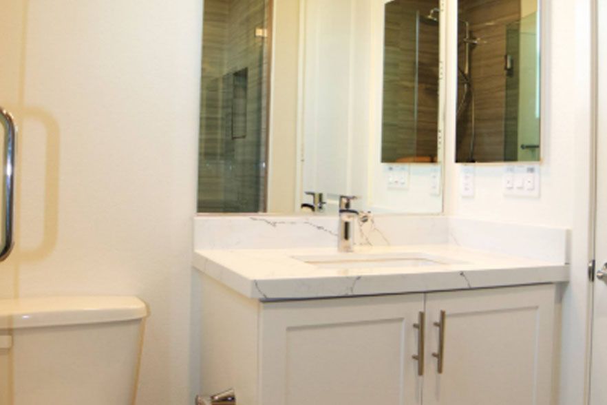White bathroom vanity with sink, mirror, and shower door, shown in a bright tiled interior