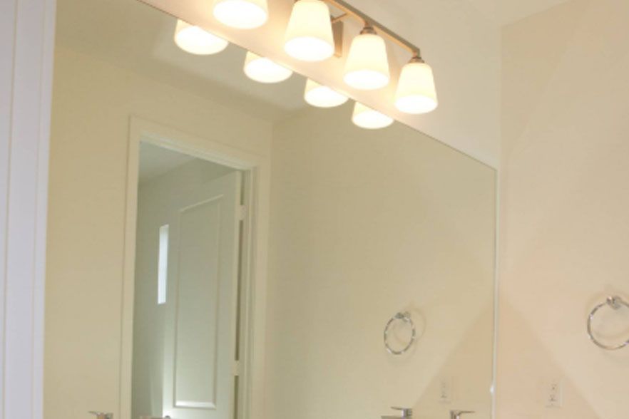 Bathroom mirror above sink with four lit vanity lights reflecting a doorway.