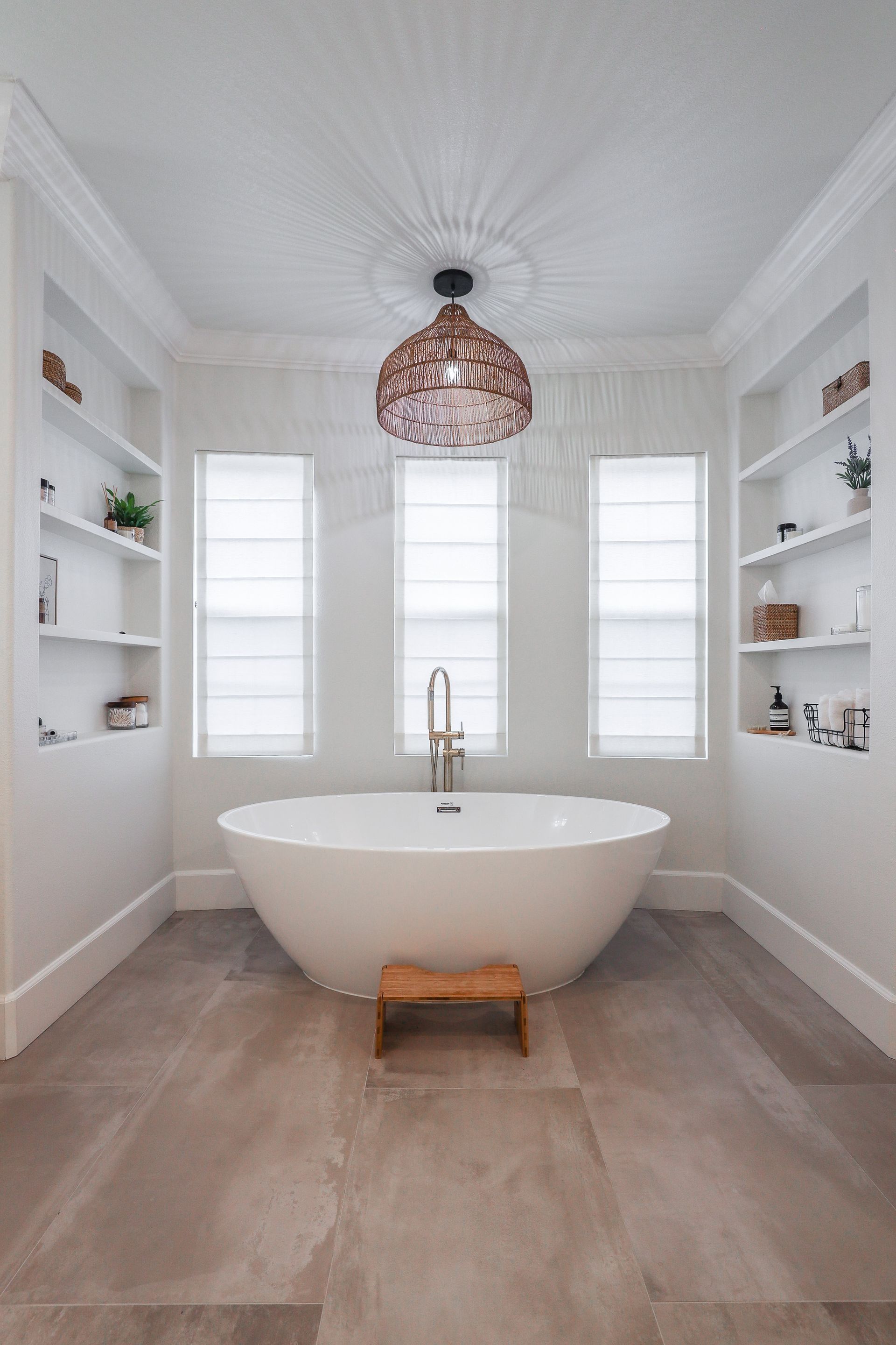 A white soaking tub sits centered under three windows in a bathroom with built-in shelves, wood-look tile, and a pendant.