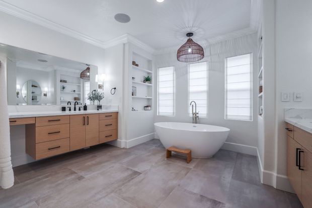 A modern, bright bathroom with light wood cabinets, a white freestanding tub, built-in shelving, and gray tile floors.